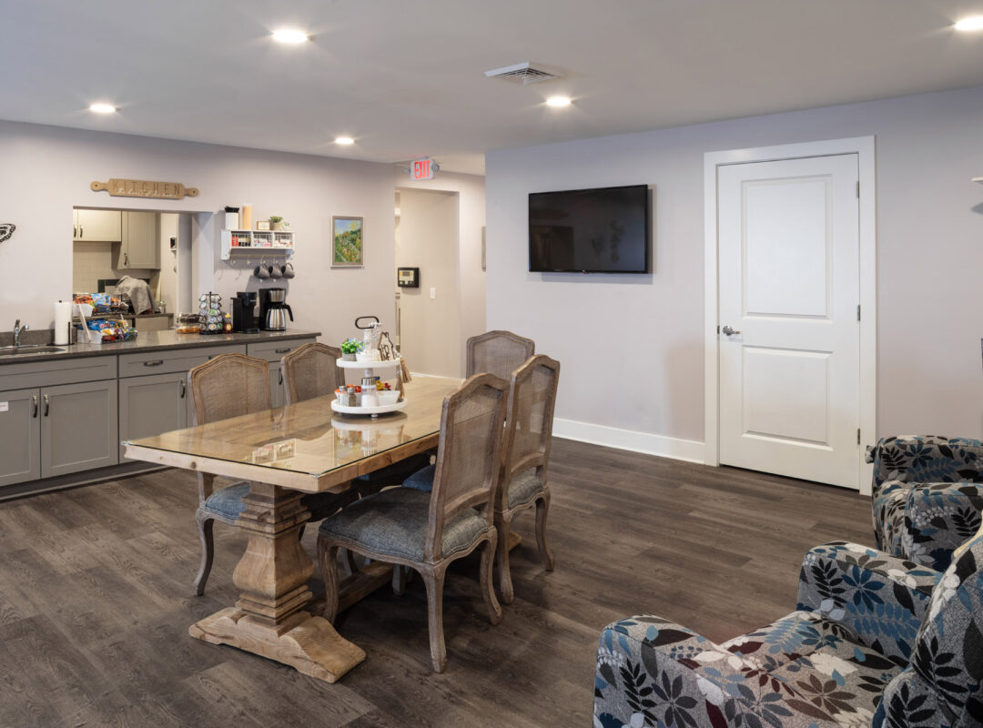 A room with a dinning table in the middle and chairs around it. There are sitting chairs and a TV on the wall.